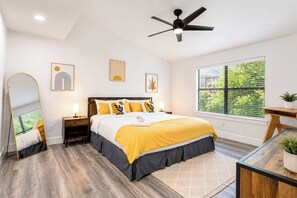 Une chambre à coucher comprenant un lit, des tables de chevet, un ventilateur de plafond, des œuvres d’art et une fenêtre donnant sur un beau paysage.