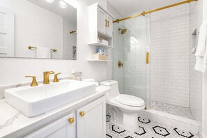 Une salle de bain avec un lavabo blanc, des robinets dorés, des toilettes, une douche en verre et un sol carrelé.
