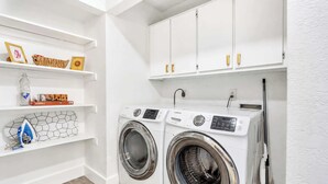 A laundry room with two front-loading washing machines, shelves with various items, and white cabinets. - Cozy East Austin 3BR for 6, Near DT & Rodeo Austin! (Austin)