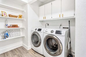 A laundry room with two front-loading washing machines, shelves with various items, and white cabinets. - Cozy East Austin 3BR for 6, Near DT & Rodeo Austin! (Austin)