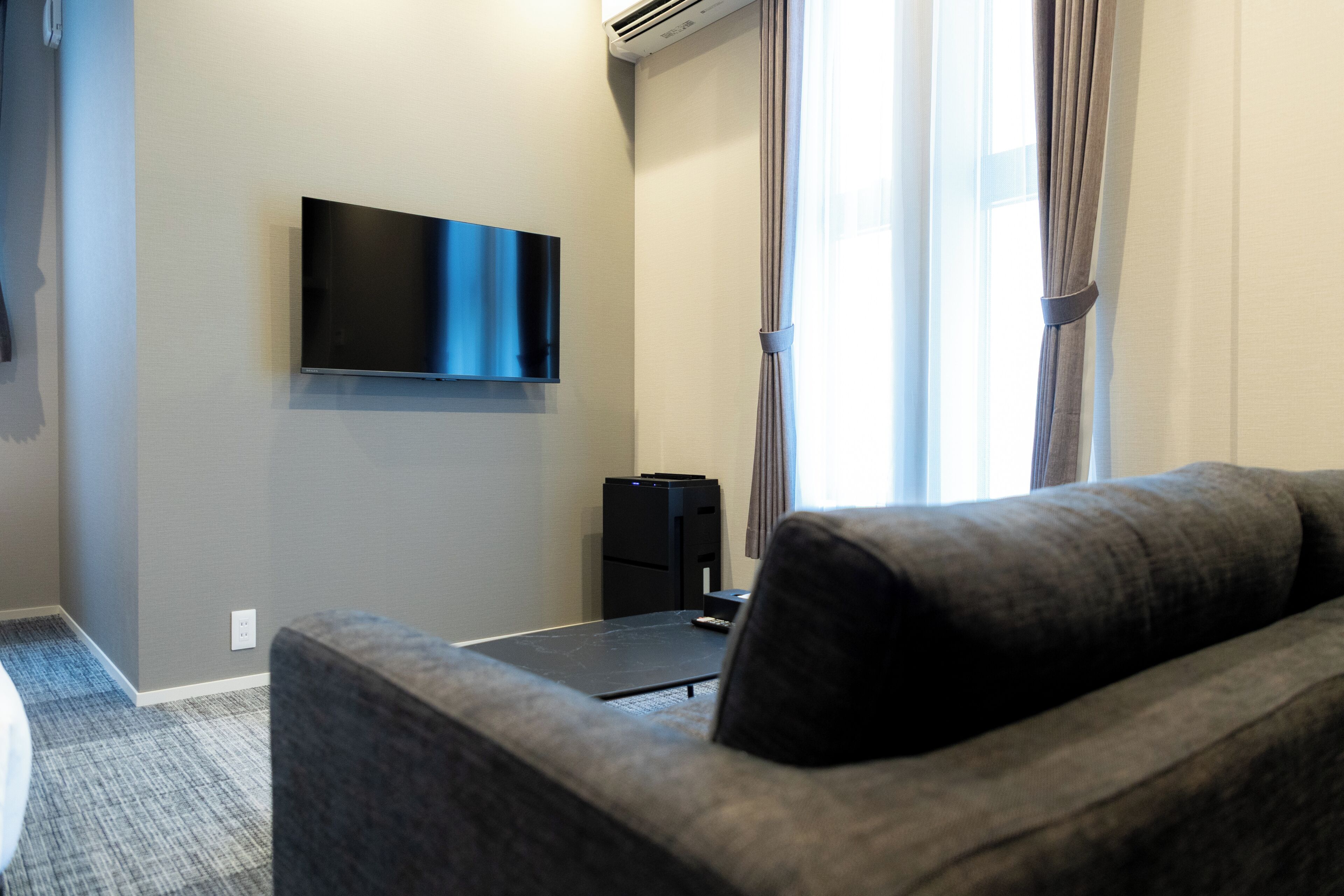 A hotel room with a flat-screen TV mounted on the wall, a grey sofa, a small black table, and a window with curtains.