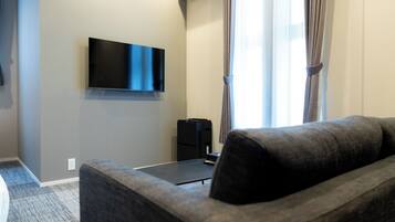 A hotel room with a flat-screen TV mounted on the wall, a grey sofa, a small black table, and a window with curtains.
