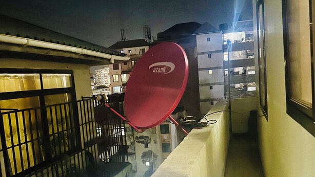 Un étroit couloir extérieur avec une antenne parabolique rouge, un mur blanc et, au fond, un bâtiment doté de nombreuses fenêtres.