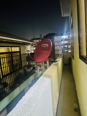 Un estrecho pasillo exterior con una antena parabólica roja, una pared blanca y un edificio con múltiples ventanas al fondo.