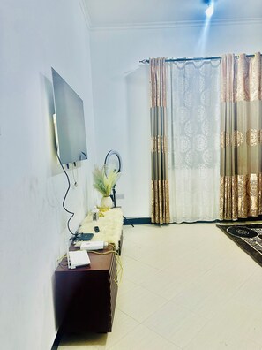 A room with a flat-screen TV mounted on the wall, a small table with a decorative vase, a power strip, and a rug on the floor. - Prime Kariakoo–Msimbazi location near markets, transport & businesses. (Dar es Salaam)