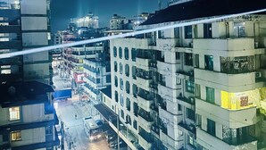 Une vue nocturne d’une ville, avec des immeubles à plusieurs étages, une rue bordée de voitures garées et un piéton visible.
