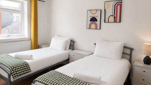 Una habitació d'hotel amb dos llits, una finestra amb cortines grogues i obra d'art a la paret.
