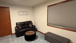 Une pièce avec un canapé en cuir foncé, un pouf rond, un banc, une grande fenêtre avec des stores et un luminaire au plafond.