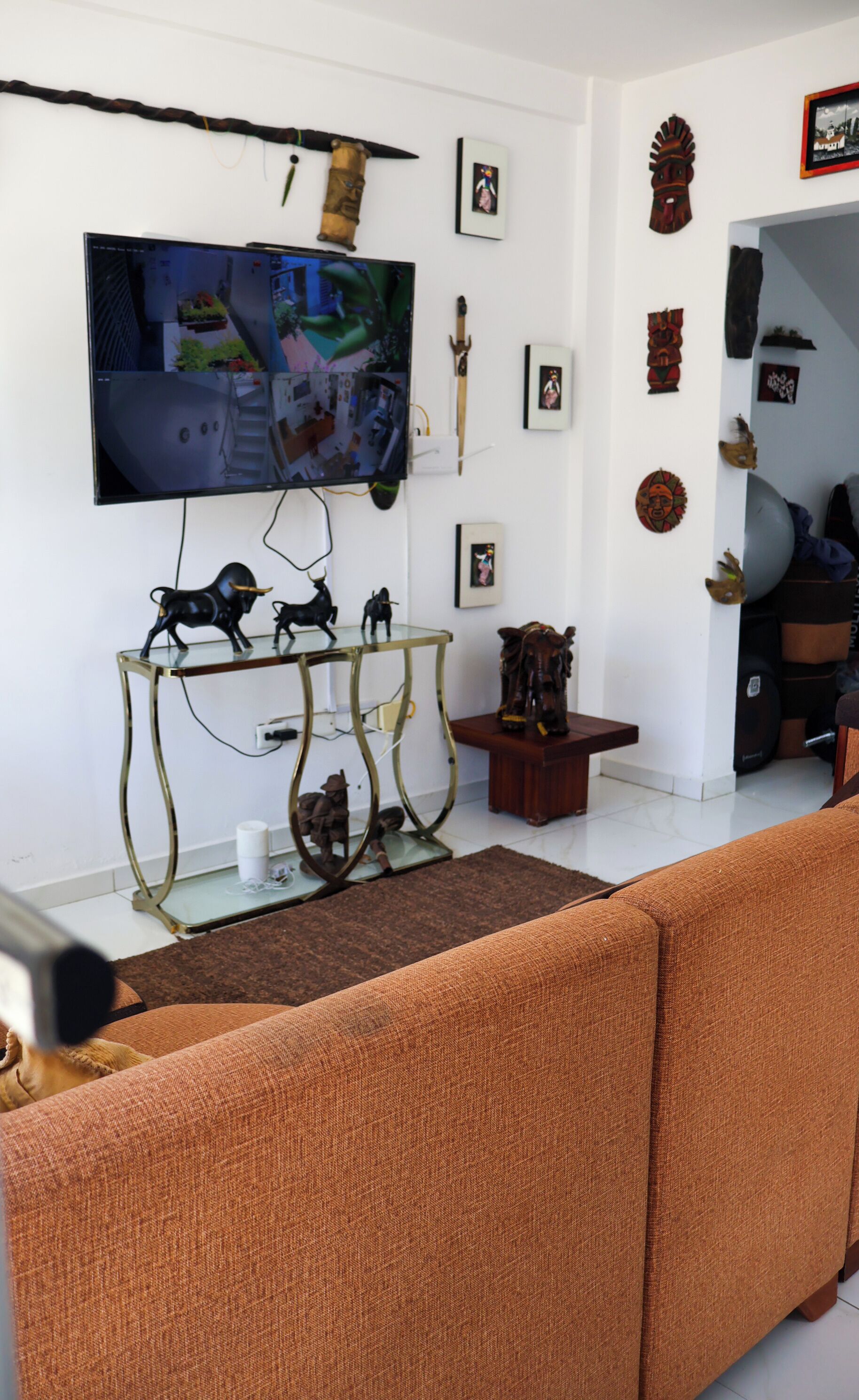 Una sala de estar con un televisor montado en la pared, una mesa de centro de vidrio con figuritas, un sofá y elementos decorativos en las paredes.
