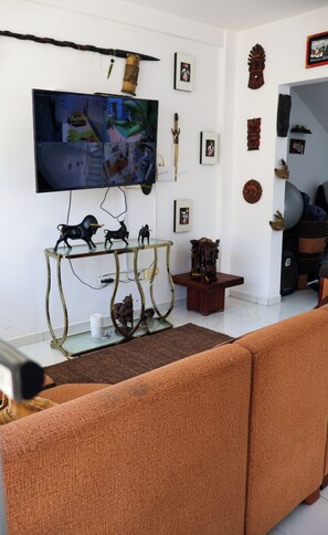 Una sala de estar con un televisor montado en la pared, una mesa de centro de vidrio con figuritas, un sofá y elementos decorativos en las paredes.