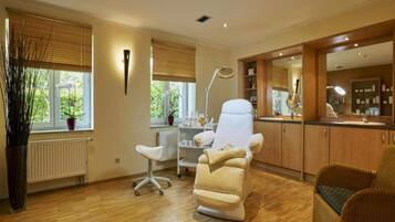 Een kamer met een massagestoel, een bureau, een stoel, een radiator, een raam met zonwering en een spiegel.