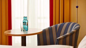 Quarto de hotel com uma poltrona listrada, uma mesa redonda com duas garrafas e copos, e um abajur.