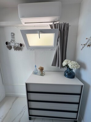 Une chambre moderne avec un bureau blanc, une petite fenêtre avec des rideaux, un climatiseur fixé au mur et un objet décoratif sur le mur.