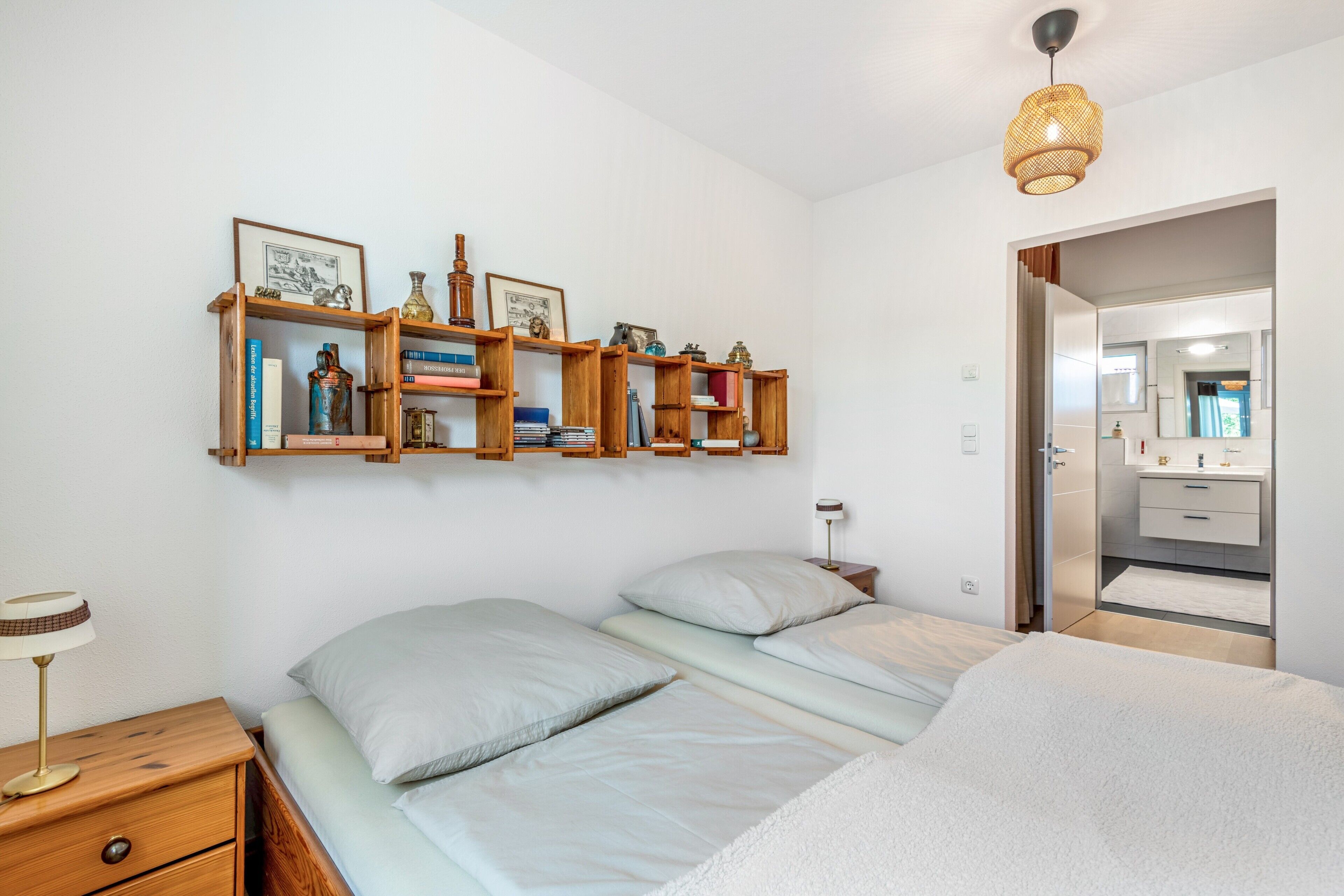 A bedroom with a bed, bedside table, lamp, and a wall-mounted shelf with books and decorative items.