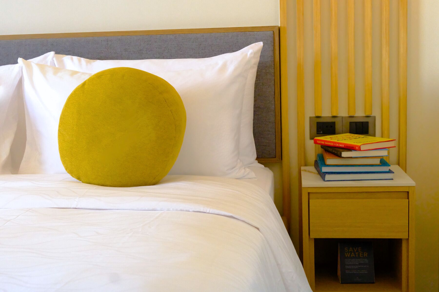 A bed with white bedding, a yellow pillow, and a bedside table with books and a 'SAVE WATER' sign.