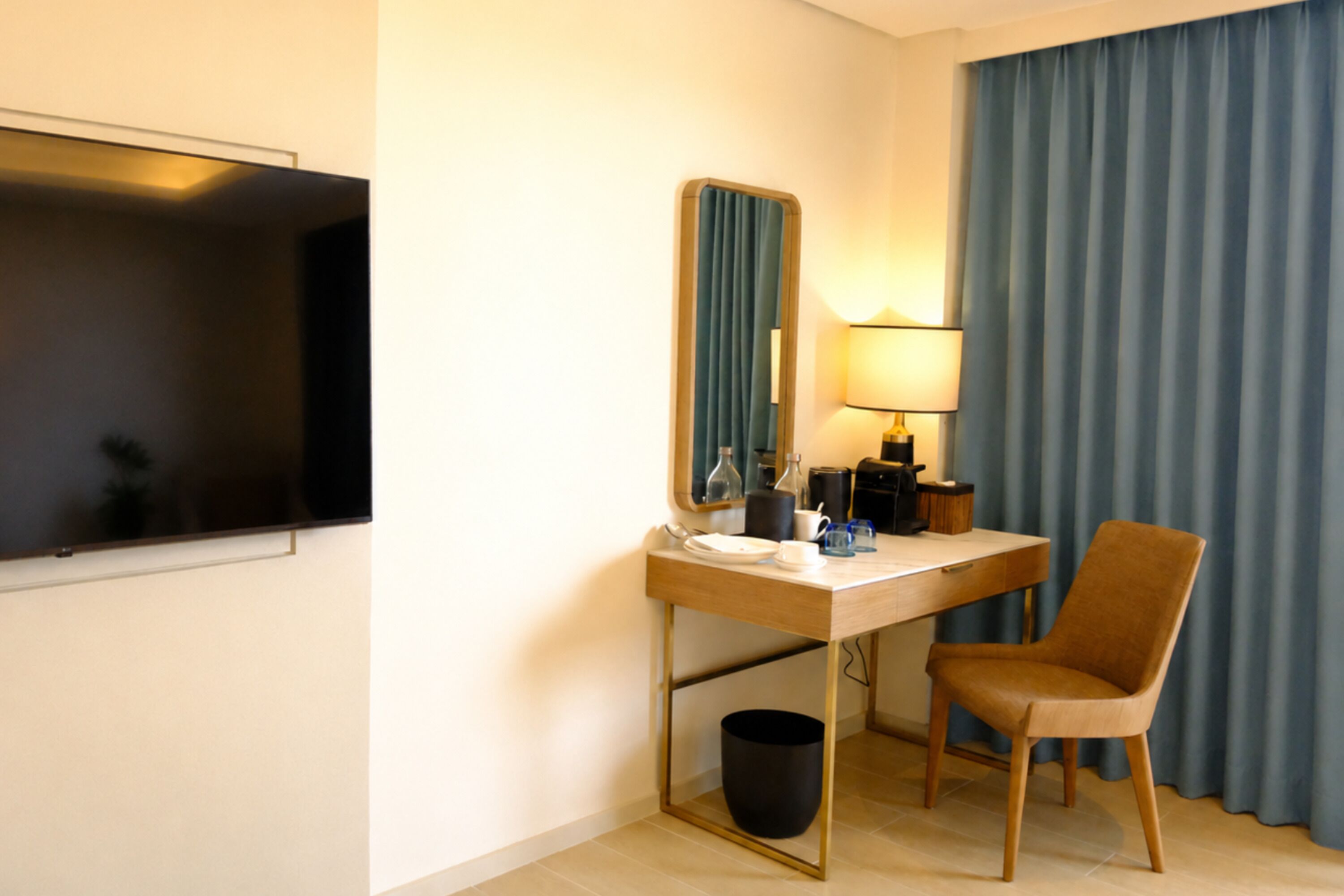 Ein Hotelzimmer mit Schreibtisch, Stuhl, Fernseher, Spiegel, Lampe und Vorhängen.