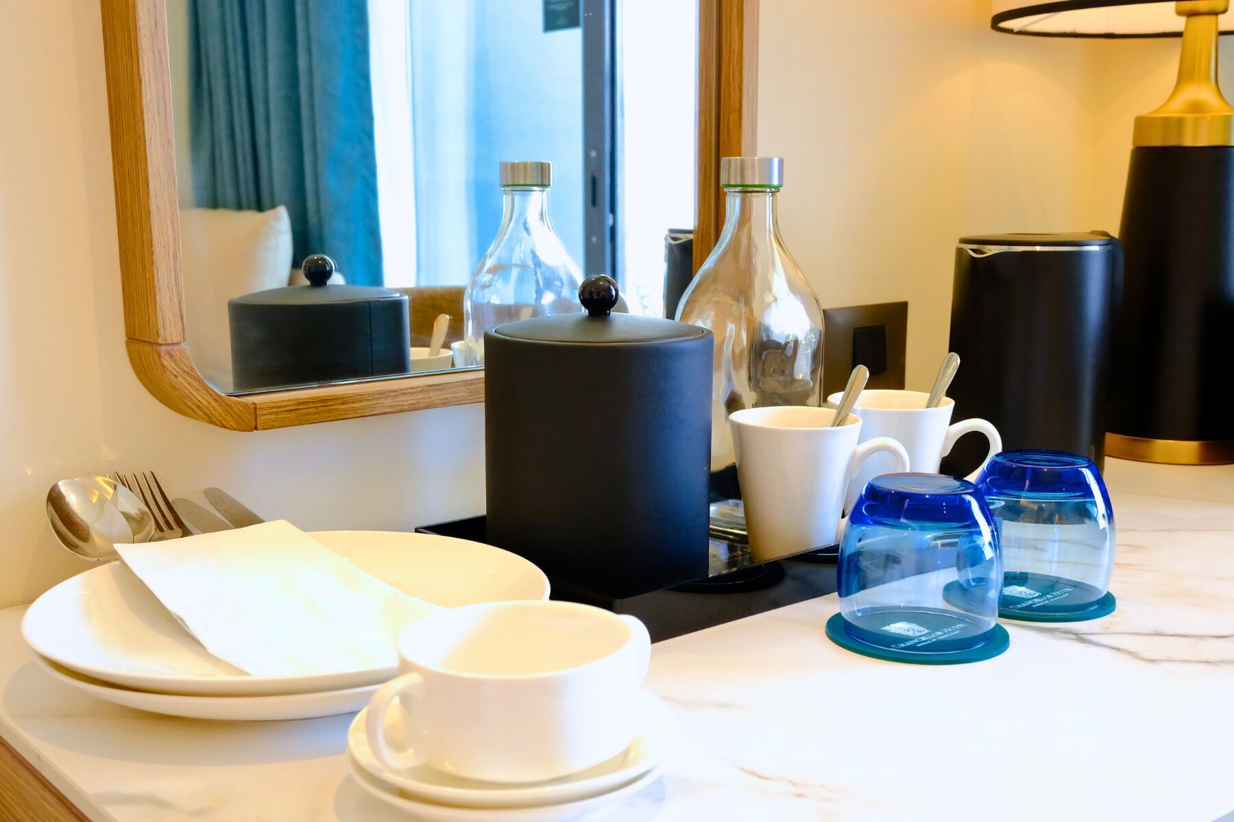 Habitación de hotel con una mesa de comedor dispuesta con platos, vasos y cubiertos. Hay un espejo en la pared, una lámpara y una botella sobre la mesa.