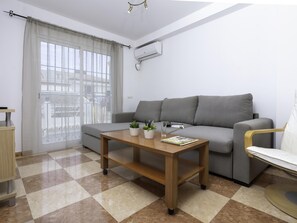 Ein modernes Wohnzimmer mit einem grauen Sofa, einem Holztisch, einem kleinen Beistelltisch und einem Sessel. Ein Fenster mit durchsichtigen Vorhängen, eine Klimaanlage an der Wand und ein Schachbrettmuster-Boden.