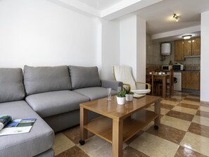 Ein Wohnzimmer mit grauem Sofa, einem Holztisch und einer Küchenzeile mit Schränken und Geräten.