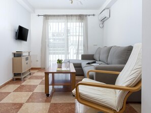 A modern living room with a sofa, a wooden chair, a small table with a plant, a flat-screen TV mounted on the wall, and a window with curtains.