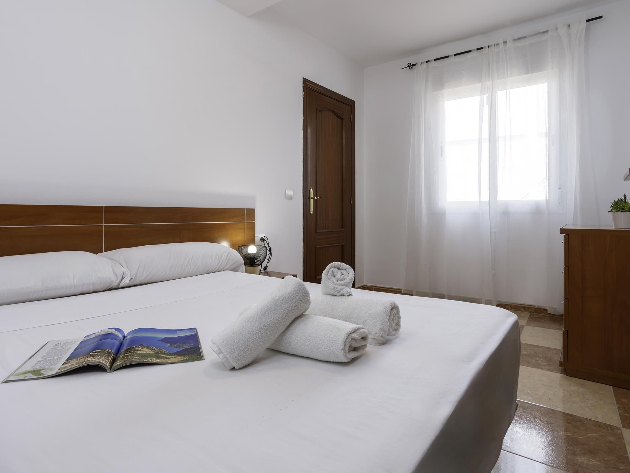 Een hotelkamer met een groot bed, een houten hoofdbord, een nachtkastje met een lamp, een deur en een raam met dunne gordijnen.