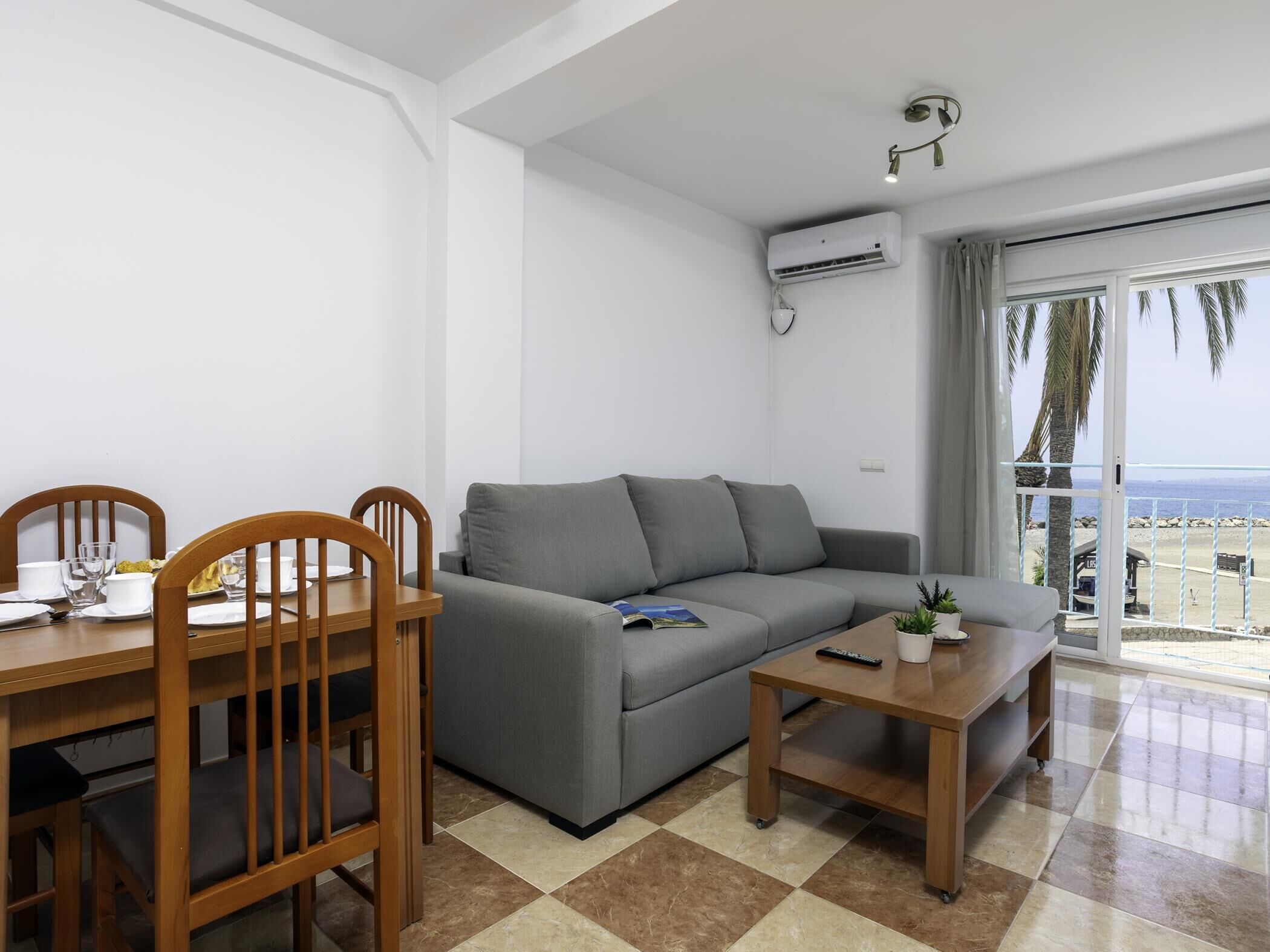 Ein modernes Wohnzimmer mit einem grauen Sofa, einem Holzespeisetisch und Stühlen. Auf einem kleinen Couchtisch liegen eine Fernbedienung und eine Pflanze.