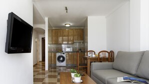 A living room with a sofa, a coffee table with a plant, a TV mounted on the wall, a kitchen with wooden cabinets, a washing machine, and a dining area with chairs and a table. - Sunstay Pedregal C Flat I Málaga (Málaga)