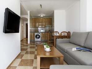A living room with a sofa, a coffee table with a plant, a TV mounted on the wall, a kitchen with wooden cabinets, a washing machine, and a dining area with chairs and a table. - Sunstay Pedregal C Flat I Málaga (Málaga)