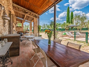 Un patio al aire libre con paredes de piedra rústica, mesas y sillas de madera, una parrilla y vistas a un jardín cercado con árboles y un cielo despejado.