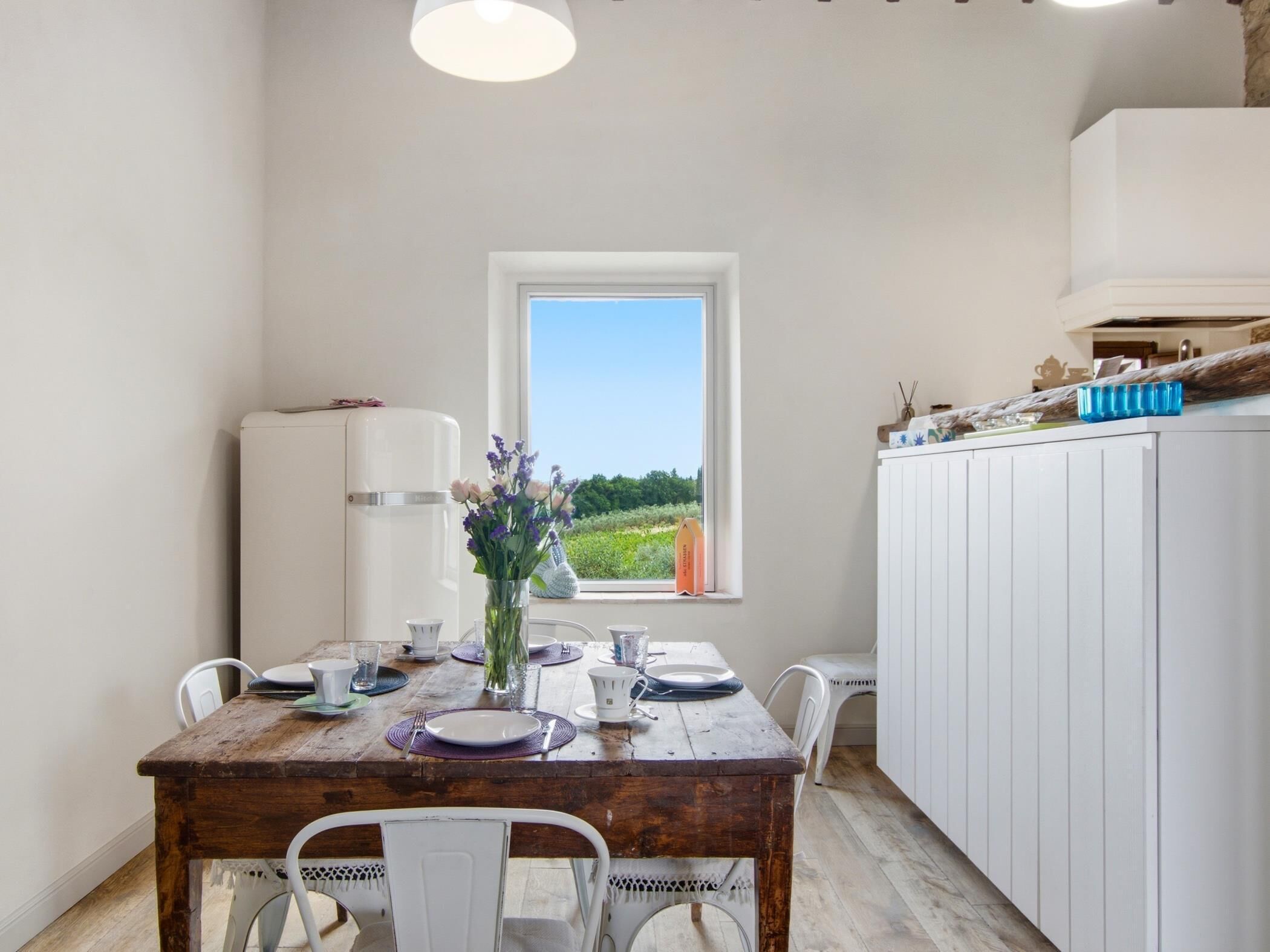 A dining area with a rustic wooden table, white chairs, a white refrigerator, and a window with a view of greenery.
