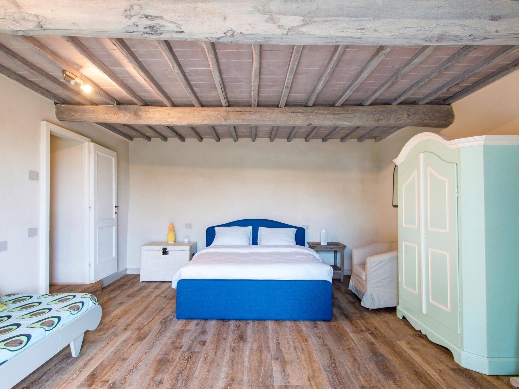 A bedroom with a wooden ceiling, a bed with a blue headboard, a nightstand, a wardrobe, and a small table with a lamp.
