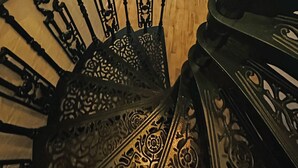 Una scala con ringhiere in metallo decorate, un pavimento in legno, un letto con materasso bianco, un termosifone e una finestra con tende.
