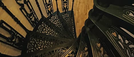 A staircase with ornate metal railings, a wooden floor, a bed with a white mattress, a radiator, and a window with curtains.