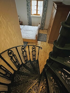 Eine Treppe mit aufwendiger Metallreling, ein Holzboden, ein Bett mit weißer Matratze, ein Heizkörper und ein Fenster mit Vorhängen.