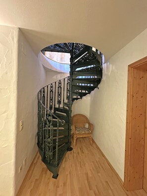 Uma escadaria em espiral com grades de metal ricamente decoradas e uma cadeira de madeira na base.