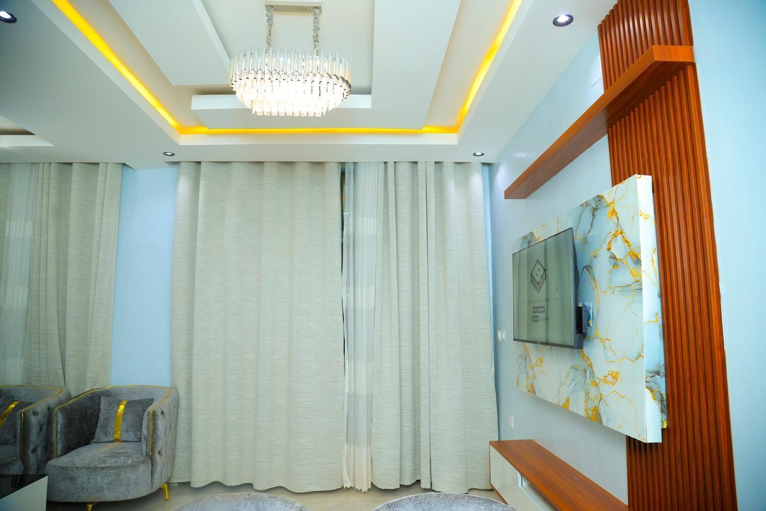 A modern living room with a chandelier, curtains, a TV mounted on a marble wall, and a grey sofa.
