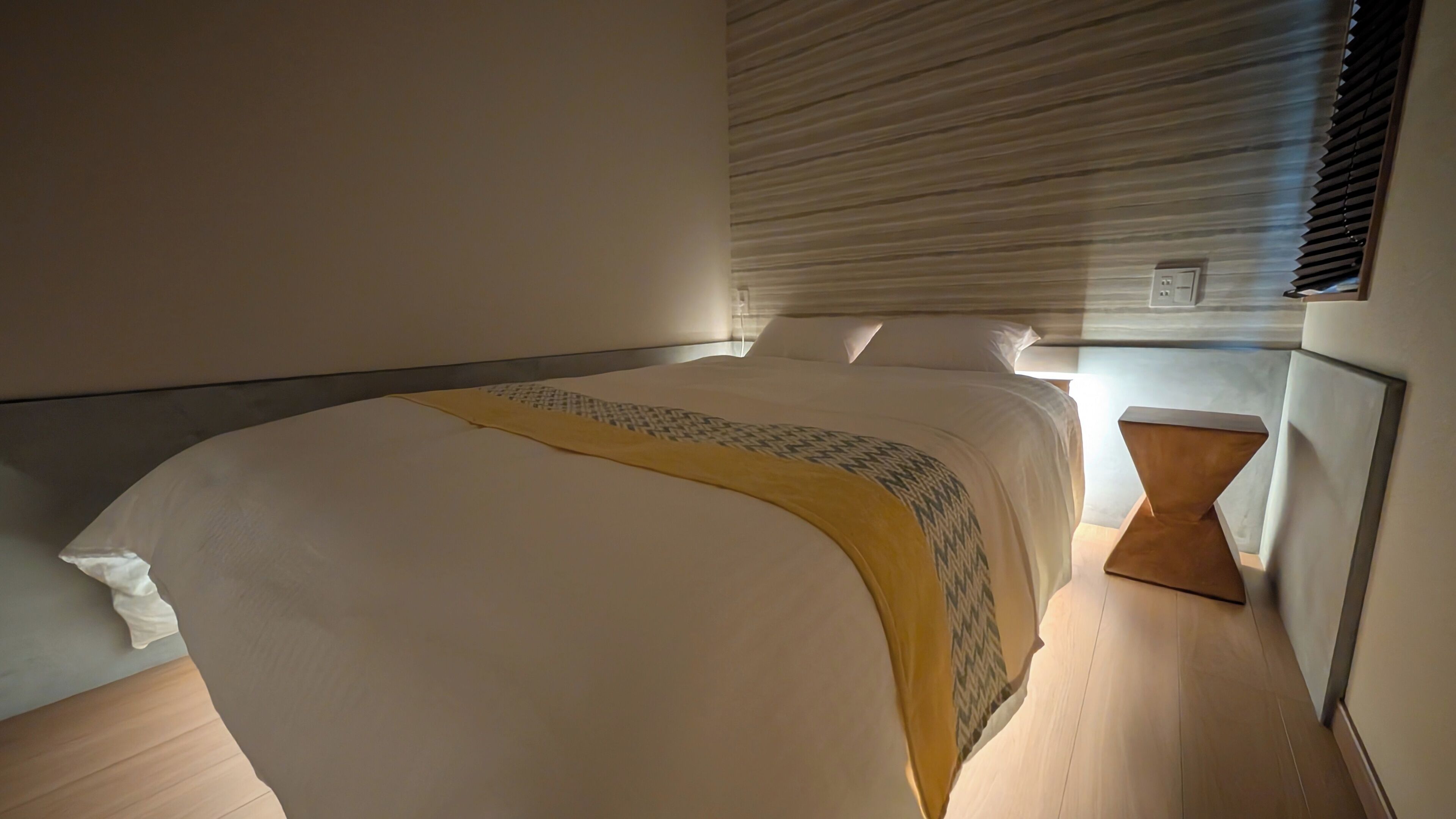 A modern bedroom with a large bed, a bedside table, and a window with blinds.