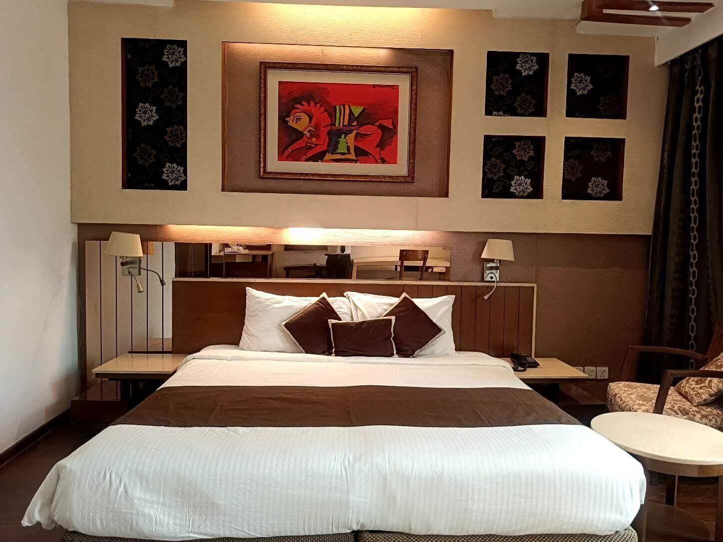 A hotel room with a large bed, a headboard with a mirror, bedside lamps, a small table, and a chair.