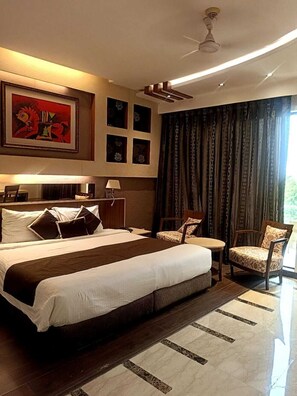 A hotel room with a large bed, a desk, two chairs, a ceiling fan, and a painting on the wall. - Club Florence (Gurugram)