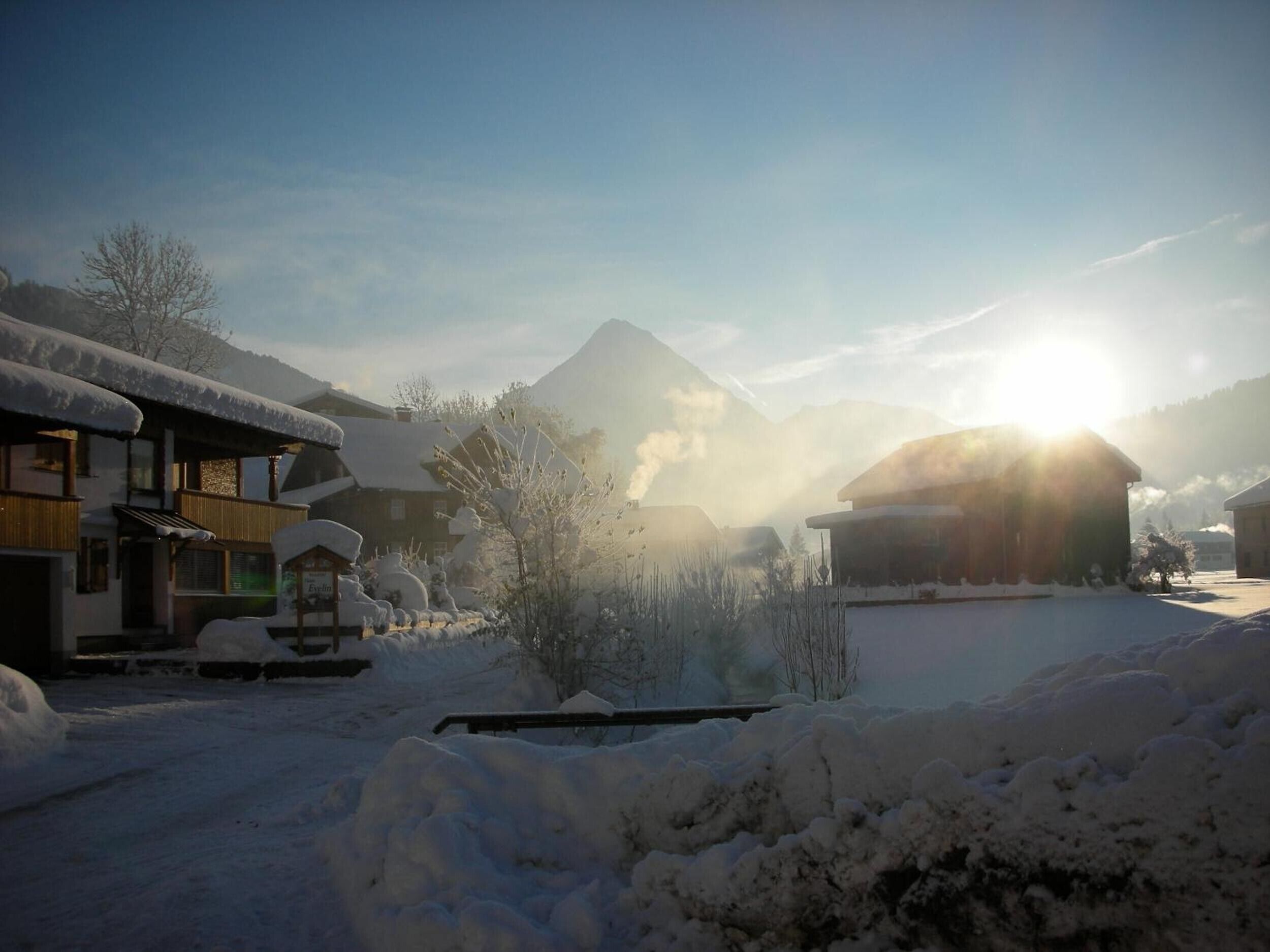 A snow-covered village with traditional buildings, a mountain in the background, and the sun shining.