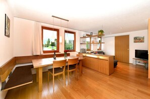 Una cocina moderna con armarios de madera, un comedor con mesa y sillas, un televisor integrado y un amplio ventanal con vistas a la vegetación.