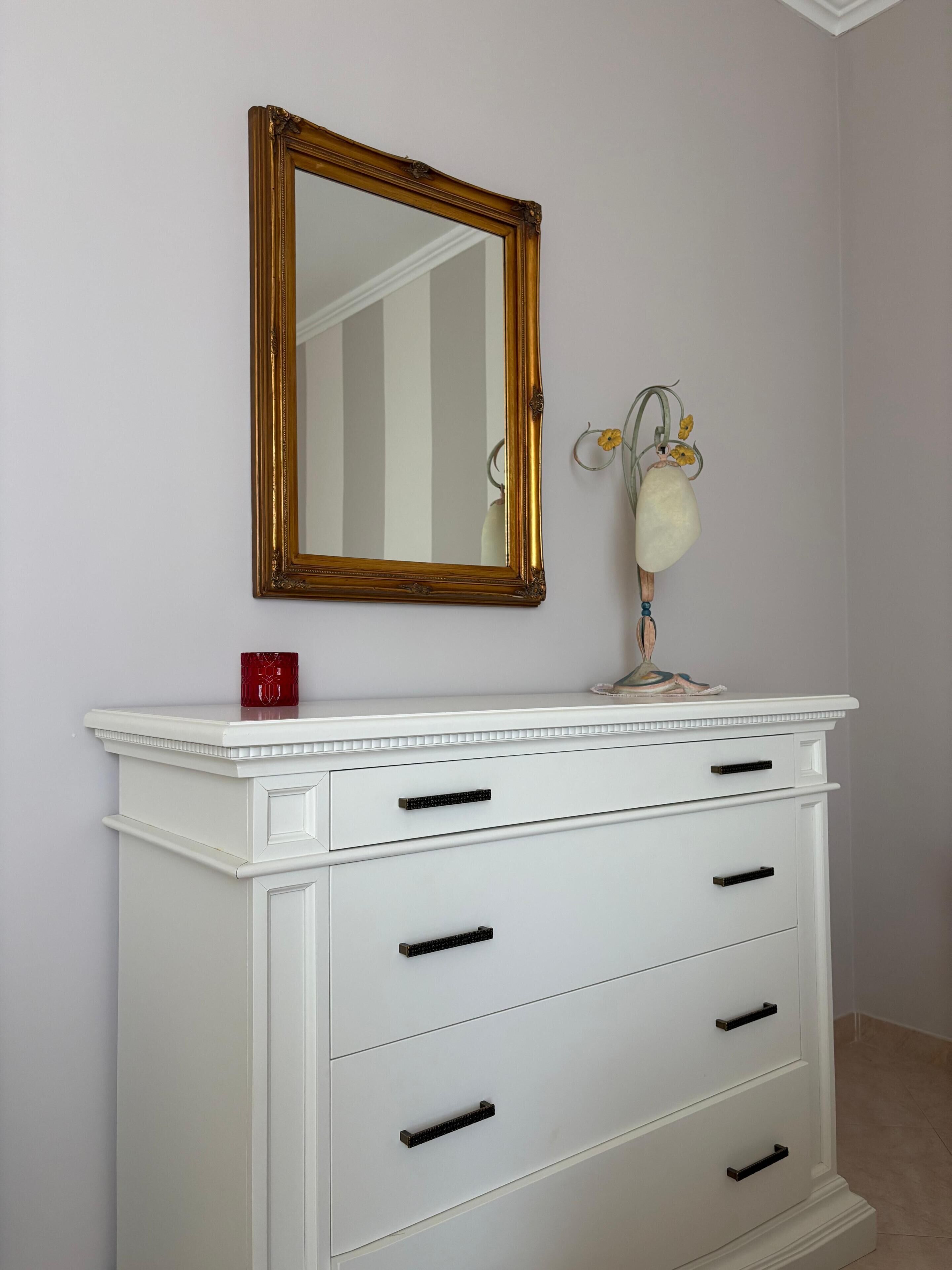 A white dresser with black handles, a gold-framed mirror above it, and a small vase with yellow flowers on the dresser's top.