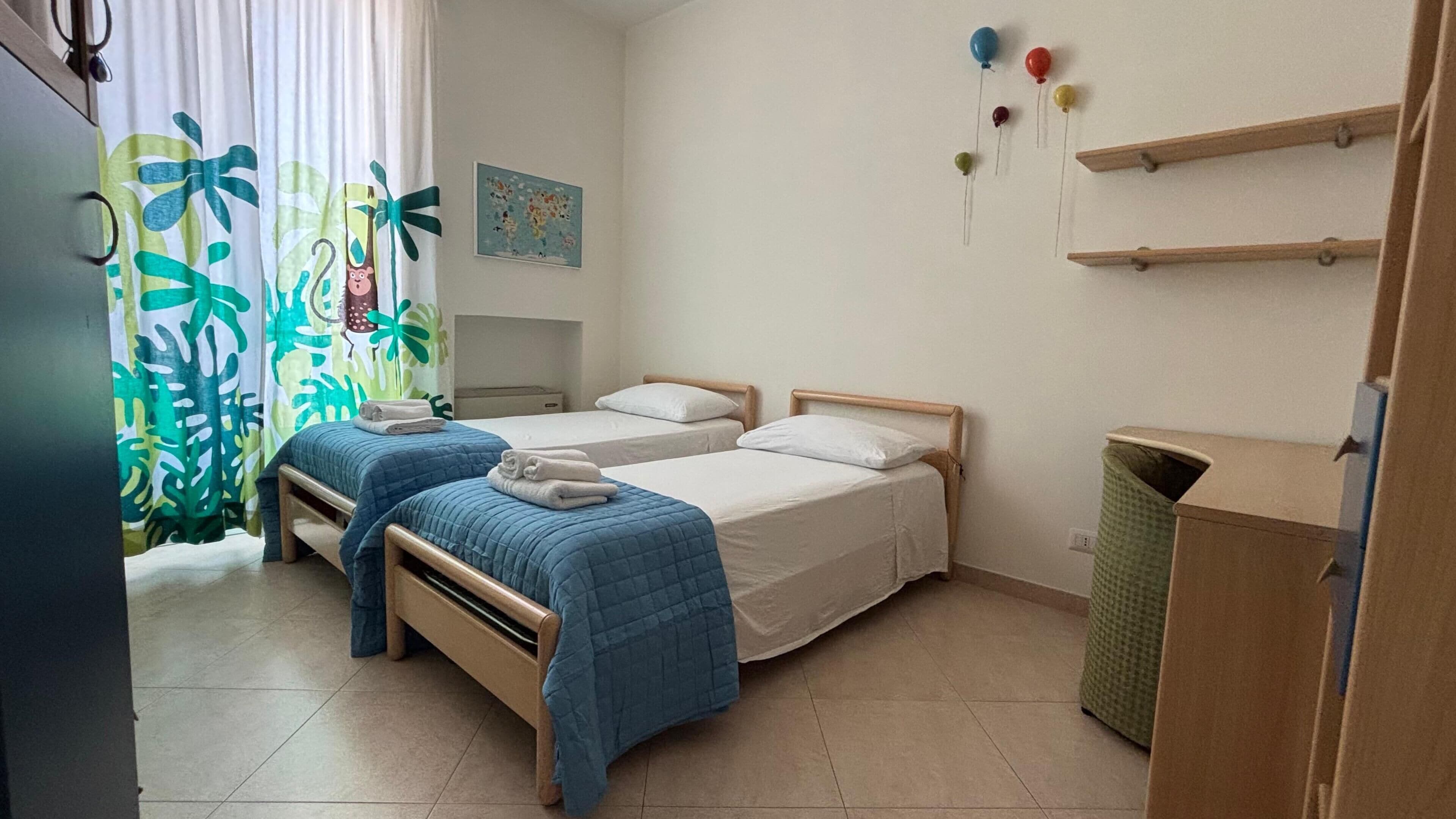 A room with two beds, a small table, a trash bin, and a wall with a picture and balloons.
