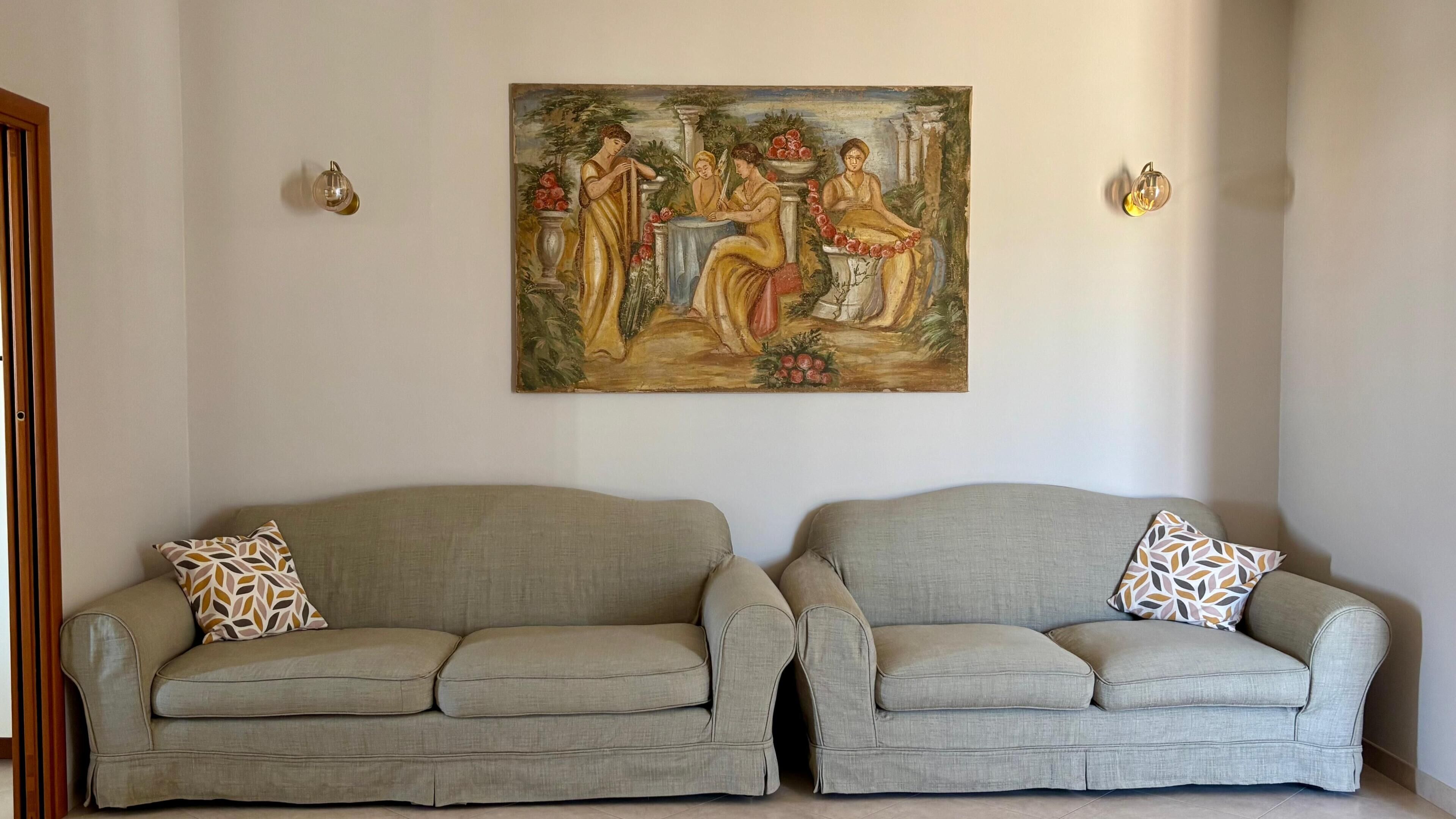 Una habitación con dos sofás grises, cada uno con cojines estampados, y un cuadro de estilo clásico en la pared.