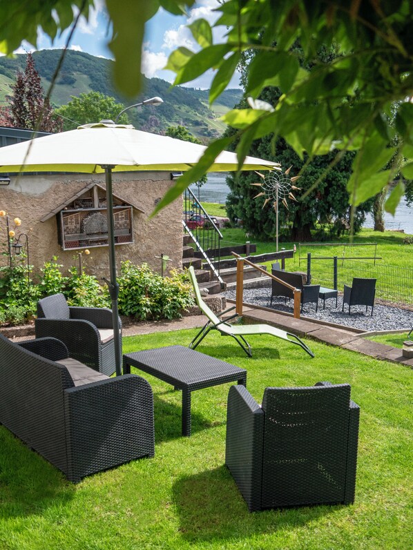 An outdoor seating area with wicker furniture, a small building with a sloped roof, and a view of a lake and hills in the background. - Hotel Gerlach (Uerzig)