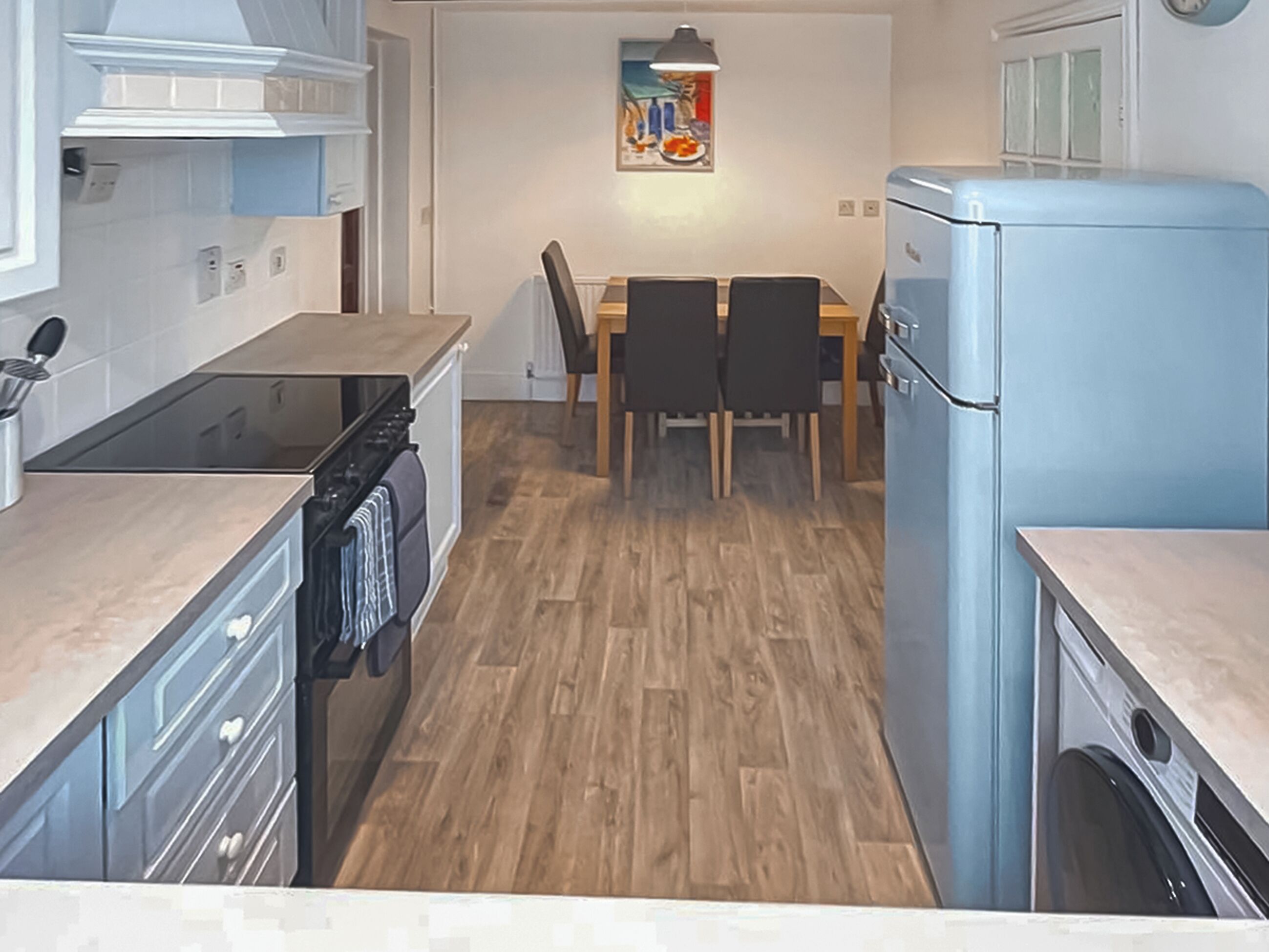 Une cuisine compacte équipée d’une cuisinière, d’un four et d’un réfrigérateur bleu. On y trouve une table à manger avec des chaises, ainsi qu’un tableau accroché au mur.