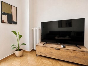 A modern living room with a large flat-screen TV, a wooden TV stand, a potted plant, and a radiator. - The Gkyzi Chapter by MPS (Athina)