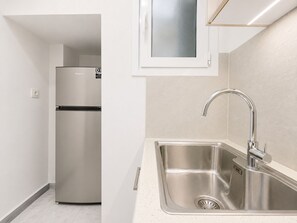 A kitchen with a stainless steel refrigerator, a sink with a modern faucet, and a window above the sink. - The Gkyzi Chapter by MPS (Athina)
