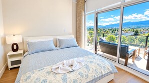 Ein Schlafzimmer mit einem großen Bett, einem Nachttisch, einer Lampe und Blick auf einen Balkon mit Gartenmöbeln.