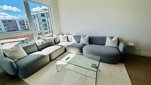 Ein modernes Wohnzimmer mit einem Ecksofa, einem Glastisch und Blick auf eine Stadtlandschaft durch große Fenster.